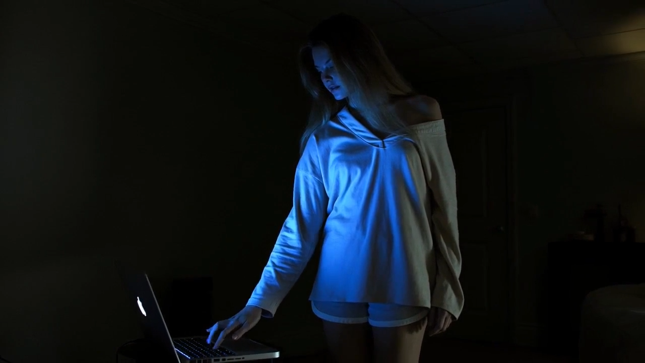Young woman in an oversized off-shoulder sweater stands in a dark room at night, lit by the blue glow of a laptop as she reaches toward the keyboard, creating a quiet and introspective mood.